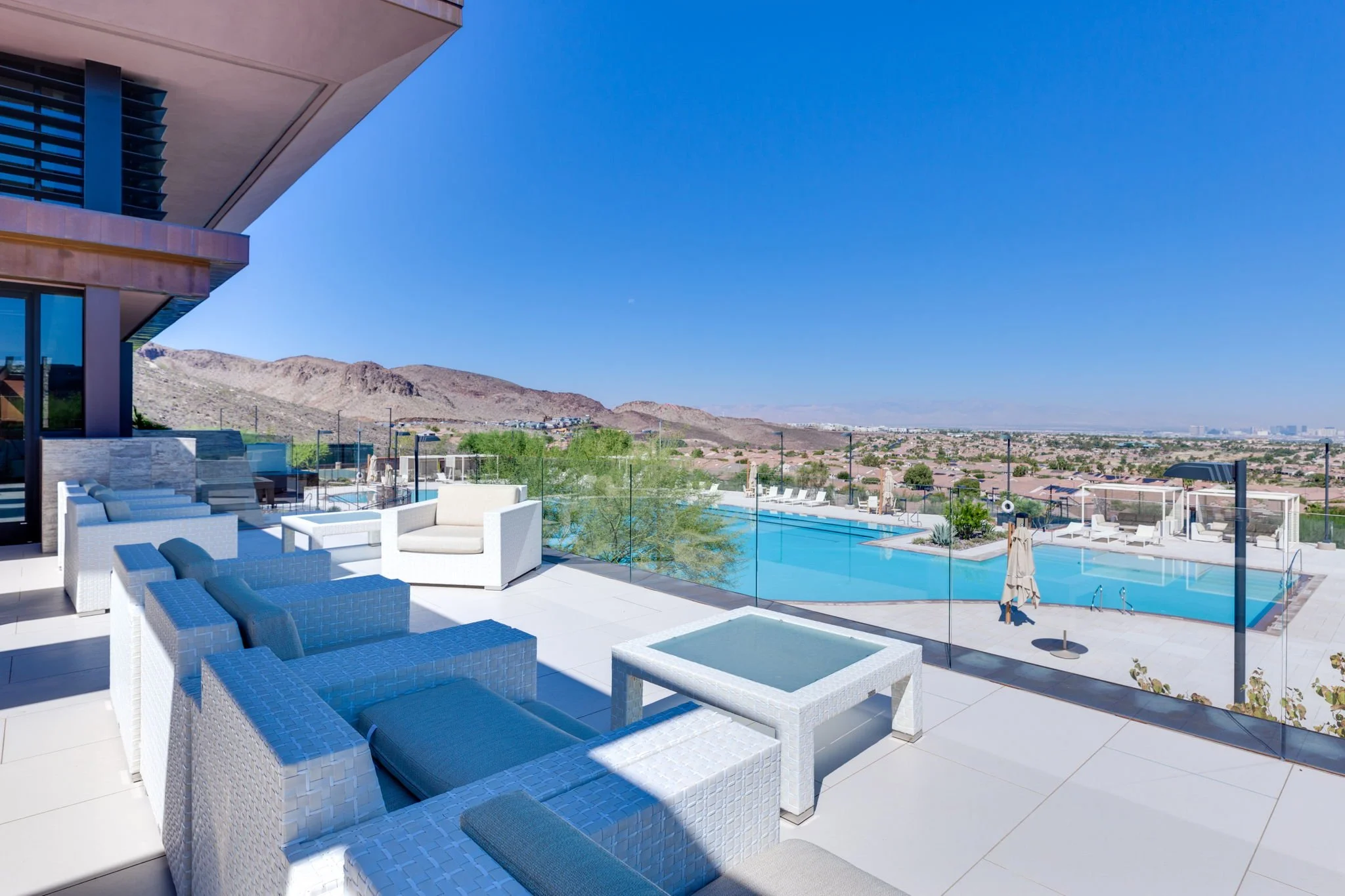 Luxury rooftop swimming pool area with modern white and blue outdoor furniture, glass railing, and scenic mountain and city views on a bright, sunny day.