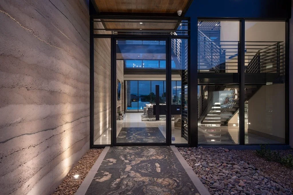Modern The Ridges entrance with stone walkway, glass double doors, interior view of living room and staircase, with a mix of stone, metal, and glass materials.