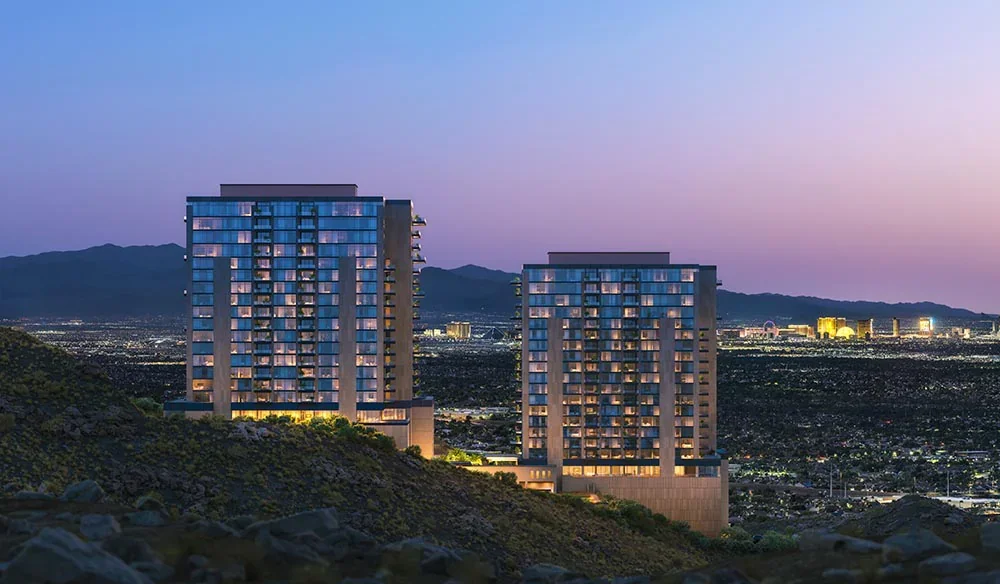 Two tall modern apartment buildings on a hillside with a cityscape and mountains in the background during twilight.
