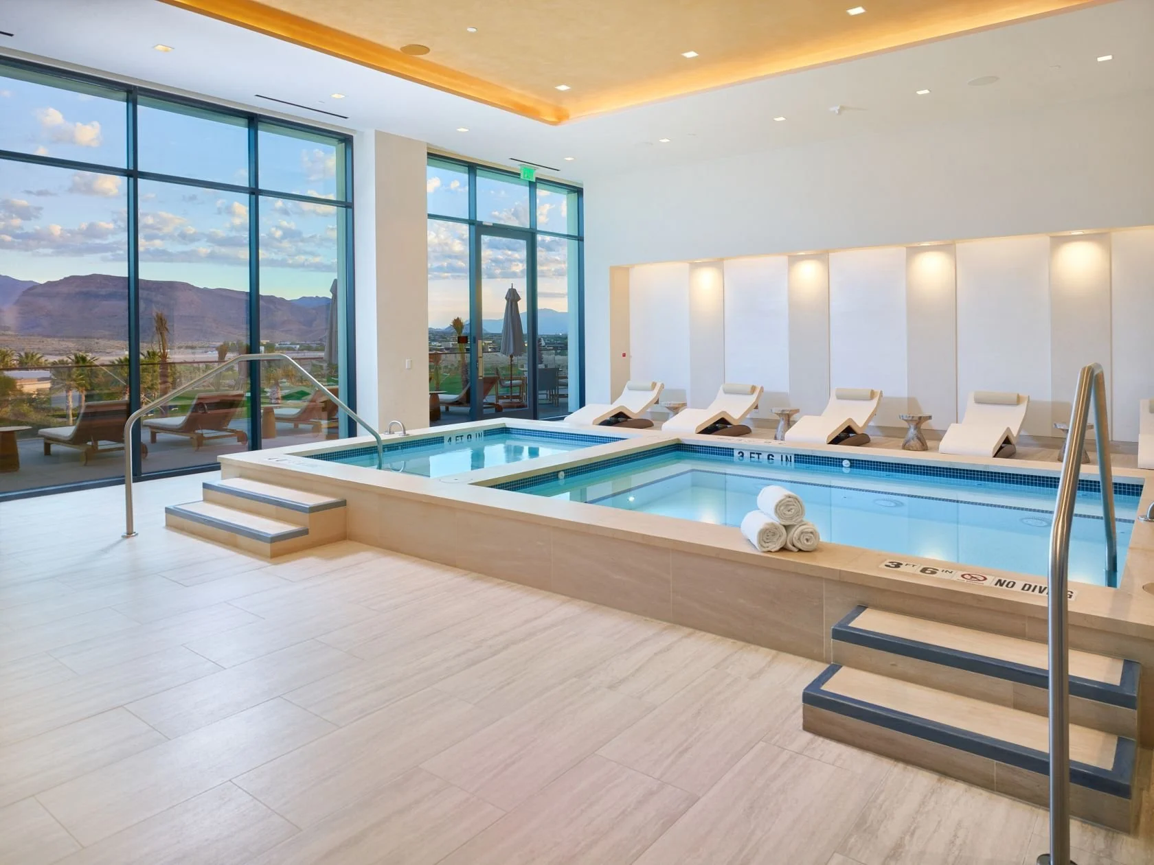Indoor swimming pool area with lounge chairs, rolled white towels, large glass windows showing mountains and blue sky, and a scenic outdoor view.