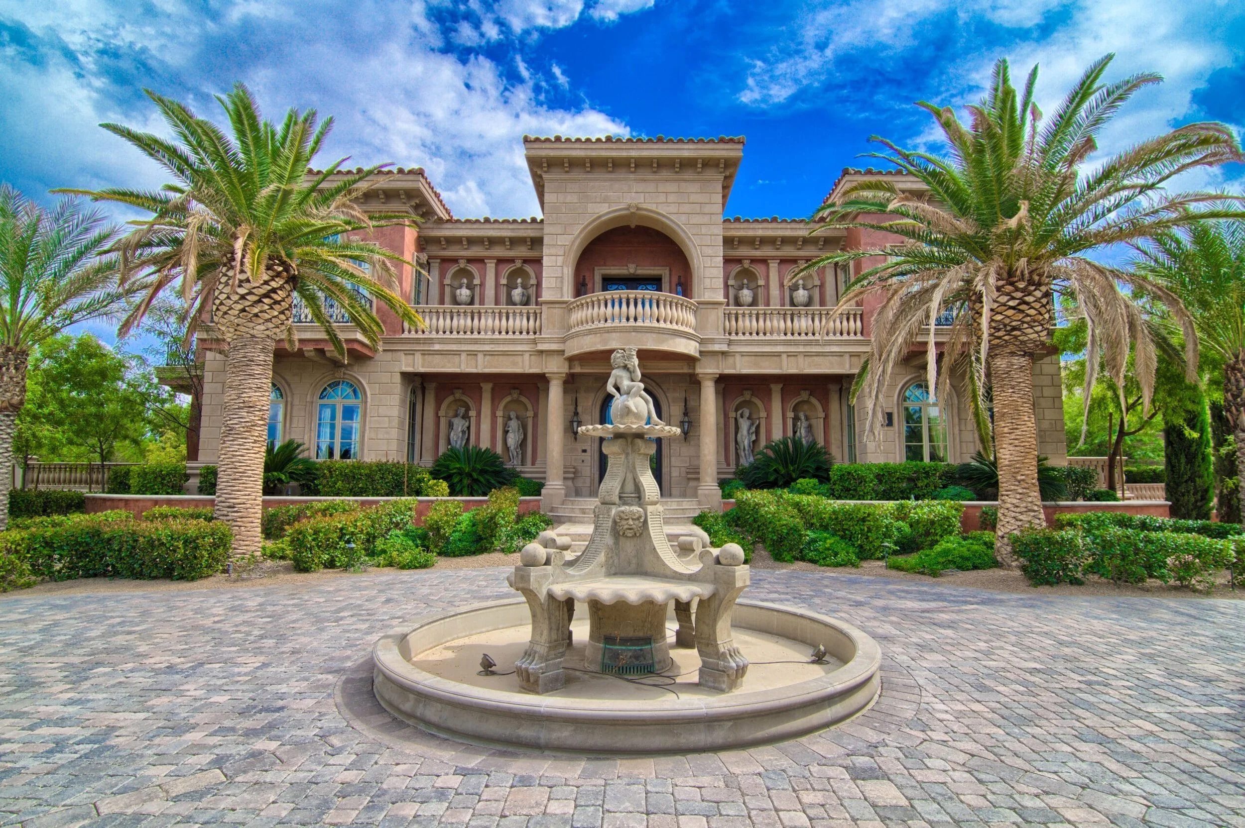 Luxury Southern Highlands Golf Club mansion with palm trees, a stone fountain, and ornate architectural details under a partly cloudy sky.