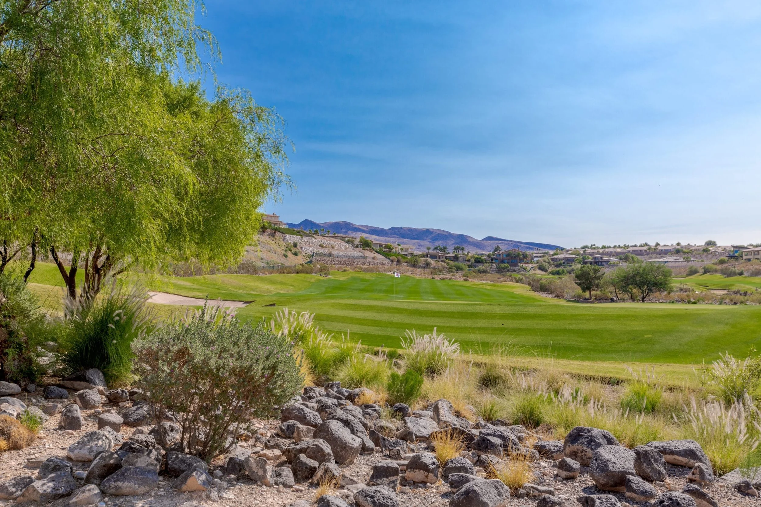 Rio Secco Golf course, Seven Hills, Henderson, with lush green fairways, surrounded by desert plants, rocks, and trees, under a clear blue sky with mountains in the background.