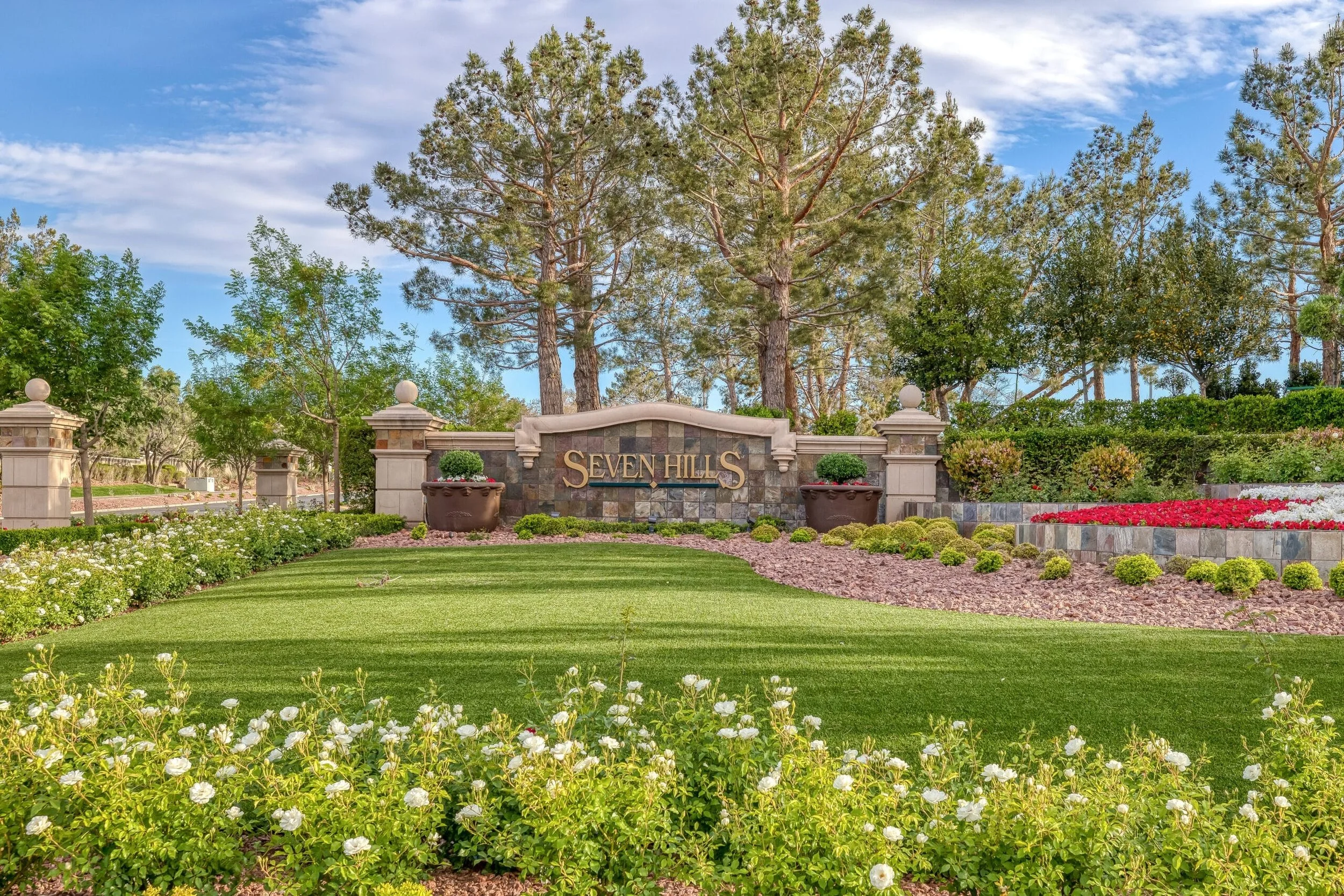 A gated entrance sign reading "Seven Hills" surrounded by well-maintained garden with green grass, flowering bushes, trees, and potted plants, set under a partly cloudy sky.