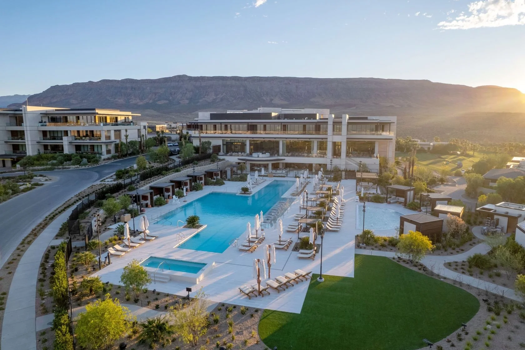 Aerial view of a modern hotel, outdoor swimming pool area with lounge chairs and umbrellas, surrounded by landscaped gardens, with mountains in the background during sunset.