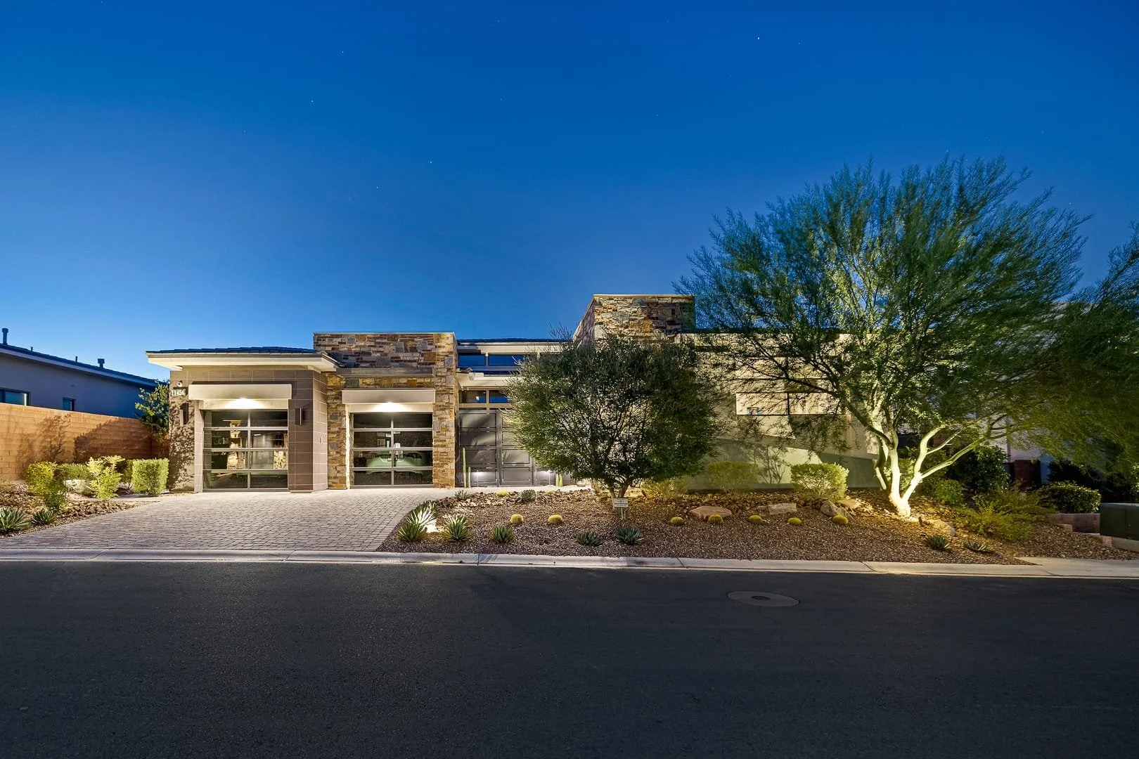 Modern house in The Ridges with a stone and beige exterior, illuminated outdoor lighting, a paved driveway, desert landscaping with small plants and trees, and a clear blue evening sky.