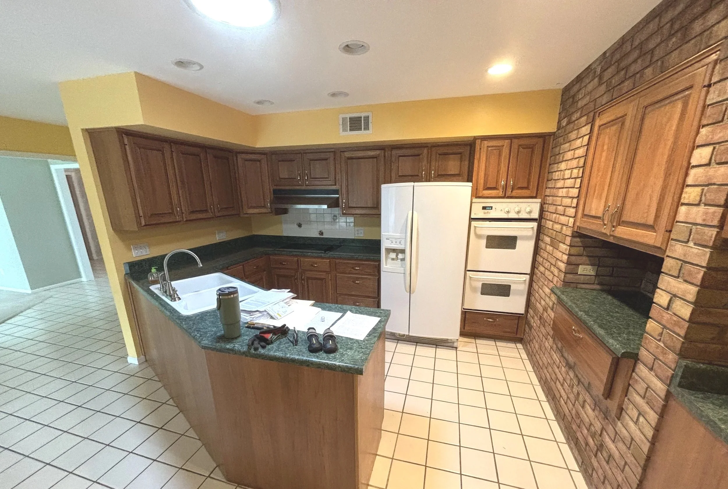 A kitchen with wooden cabinets, green granite countertops, a white refrigerator, and a brick accent wall. The kitchen island has some papers, tools, and a water bottle on it.