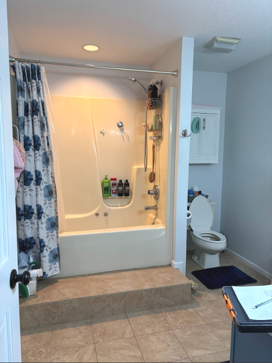Bathroom with a bathtub and shower, shower curtain with blue flowers, toiletries inside the tub, toilet, and a wall-mounted cabinet.
