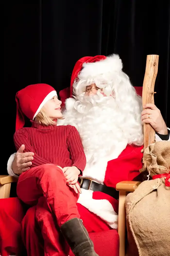 A young girl sitting on Santa Claus's lap, both wearing red Santa hats, with Santa holding a wooden staff and dressed in traditional red and white suit, sitting against a black background.