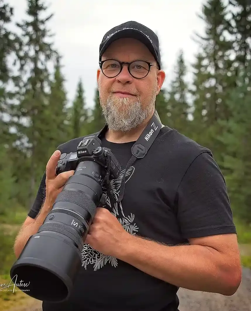 A man with glasses and a beard holding a Nikon camera with a large telephoto lens outdoors in a forested area.