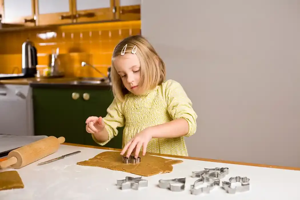 A young girl with blonde hair and hair clips, wearing a yellow dress, is using cookie cutters to cut out shapes from rolled-out gingerbread dough on a kitchen counter.
