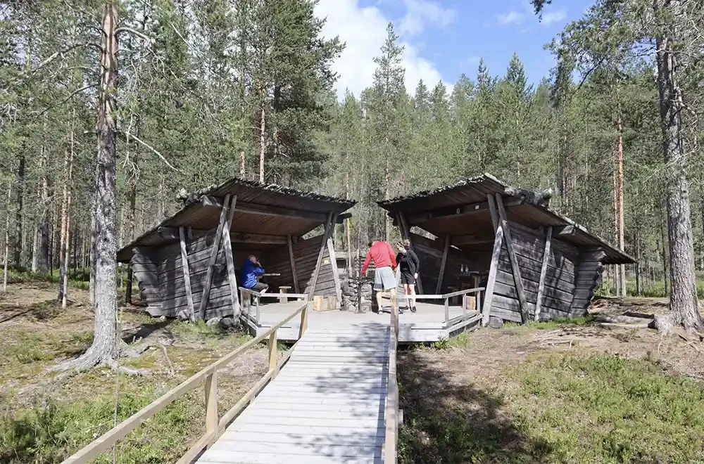 Two rustic wooden cabins in a forest, connected by a wooden walkway, with a few people walking and sitting around them.
