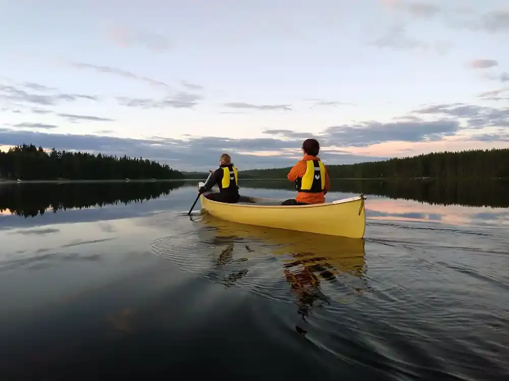 Two people in a yellow canoe paddling on a calm lake at sunset, with forested shoreline in the background.