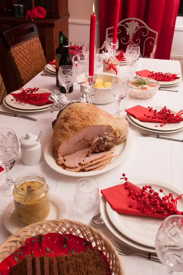 A festive holiday dinner table with a large roasted meat, salad, bread, red napkins, red berries, candles, and wine glasses.