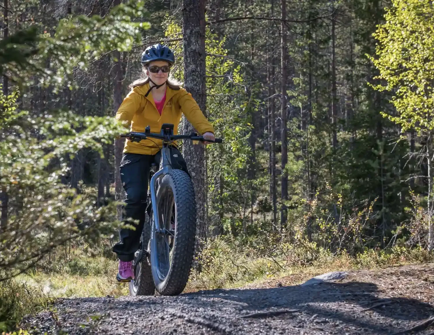 A woman wearing a yellow jacket and helmet riding a mountain bike on a dirt trail through a forest.