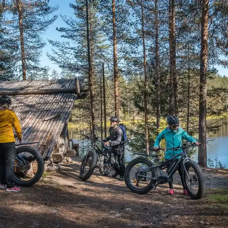 Three people with electric bikes on a forest trail by a lake, with trees and a rustic cabin in the background.