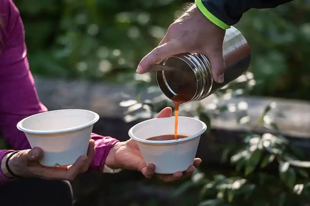 Person pouring hot coffee from a thermal flask into a paper cup held by another person outdoors.