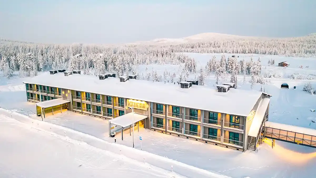 Snow-covered motel building in a winter landscape with trees and hills in the background, with warm lights glowing inside.