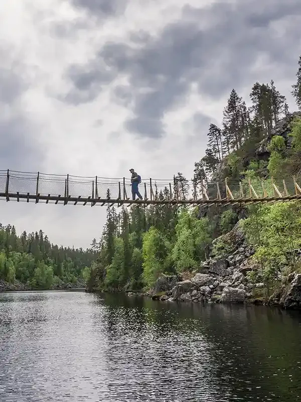 Person walking on a narrow wooden suspension bridge over a river, surrounded by green trees and rocky cliffs, under a cloudy sky.