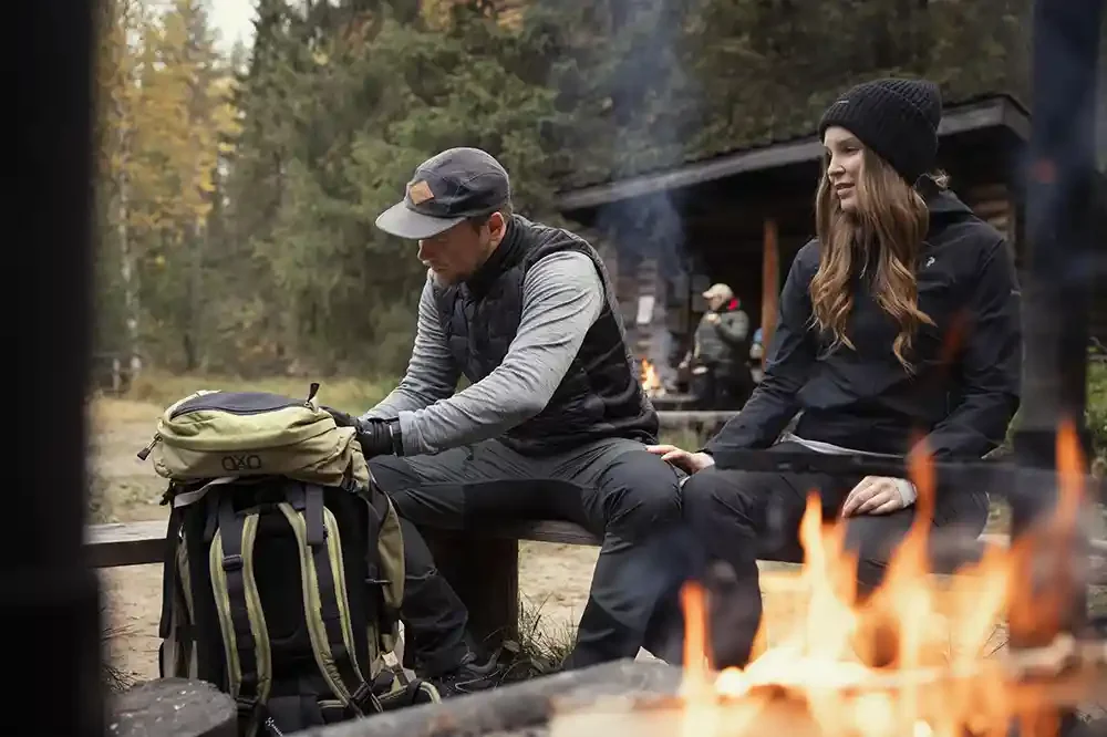 Two people sitting on a wooden bench outdoors near a campfire in a forest. One person is looking into a backpack, and the other is looking at them while smiling. The background shows trees and a small cabin.
