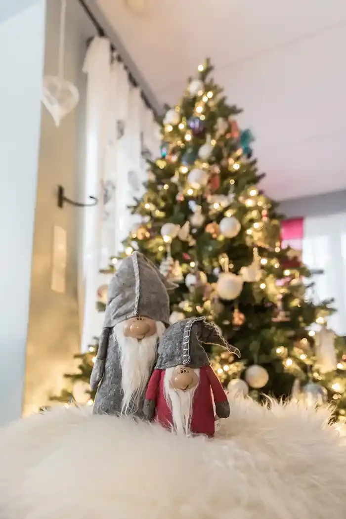Two small gnome figurines with long white beards and pointed hats, one in gray and one in red, placed on a fluffy white surface in front of a decorated Christmas tree with white ornaments and warm lights.