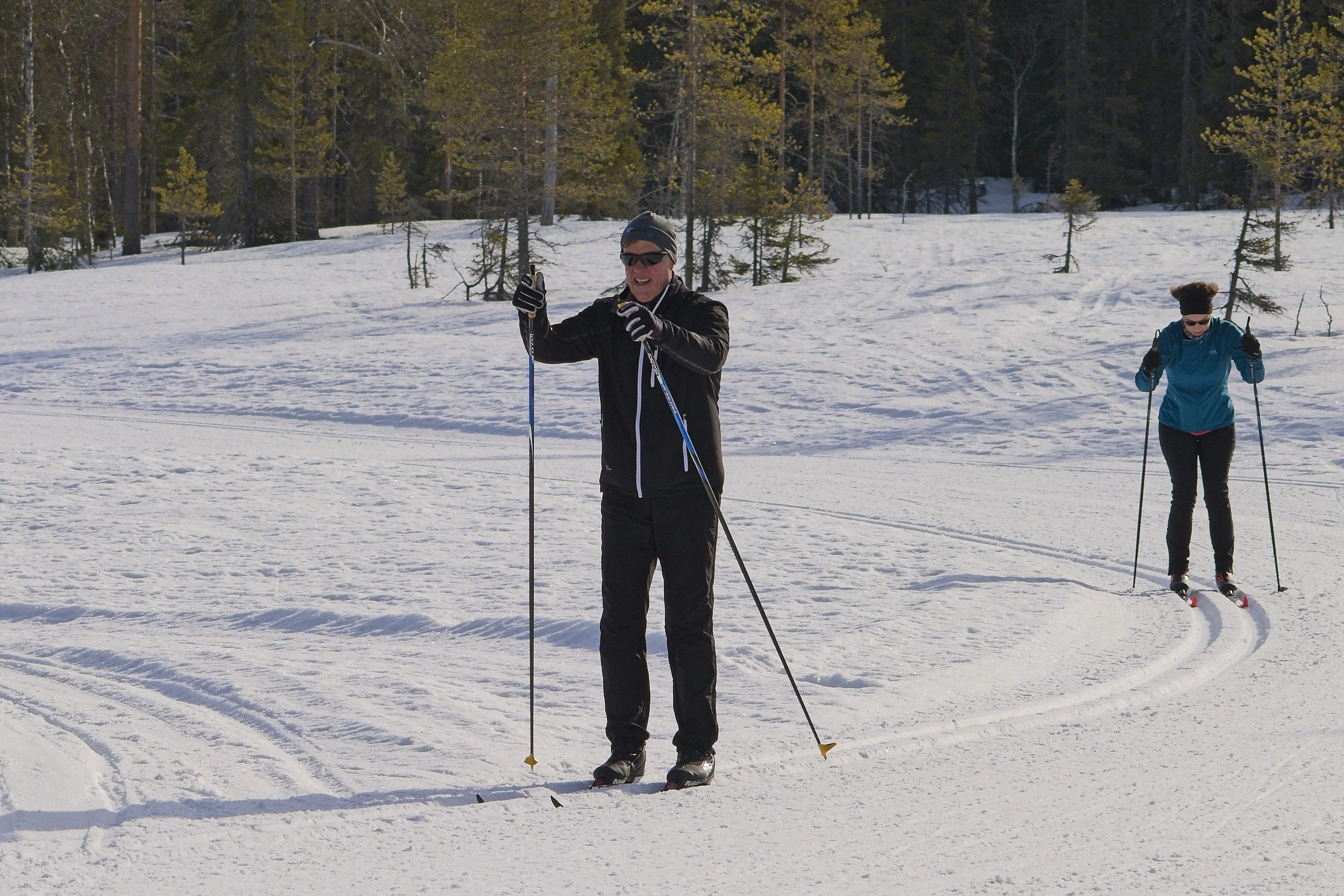 Two people cross-country skiing on snow-covered ground in a forested area with tall pine trees and a clear sky. The person in the foreground is smiling and pointing forward, while the person in the background is looking down at the snow.