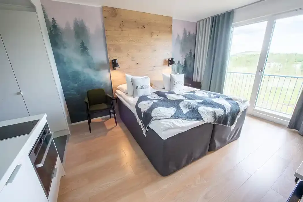 A bedroom with a large bed featuring a wooden headboard, two black wall-mounted lamps, and two pillows that say 'KID' and 'SCOOL'. There is a green armchair beside the bed, a white wardrobe, and a sliding glass door leading to a balcony with a scenic view of trees and mountains.