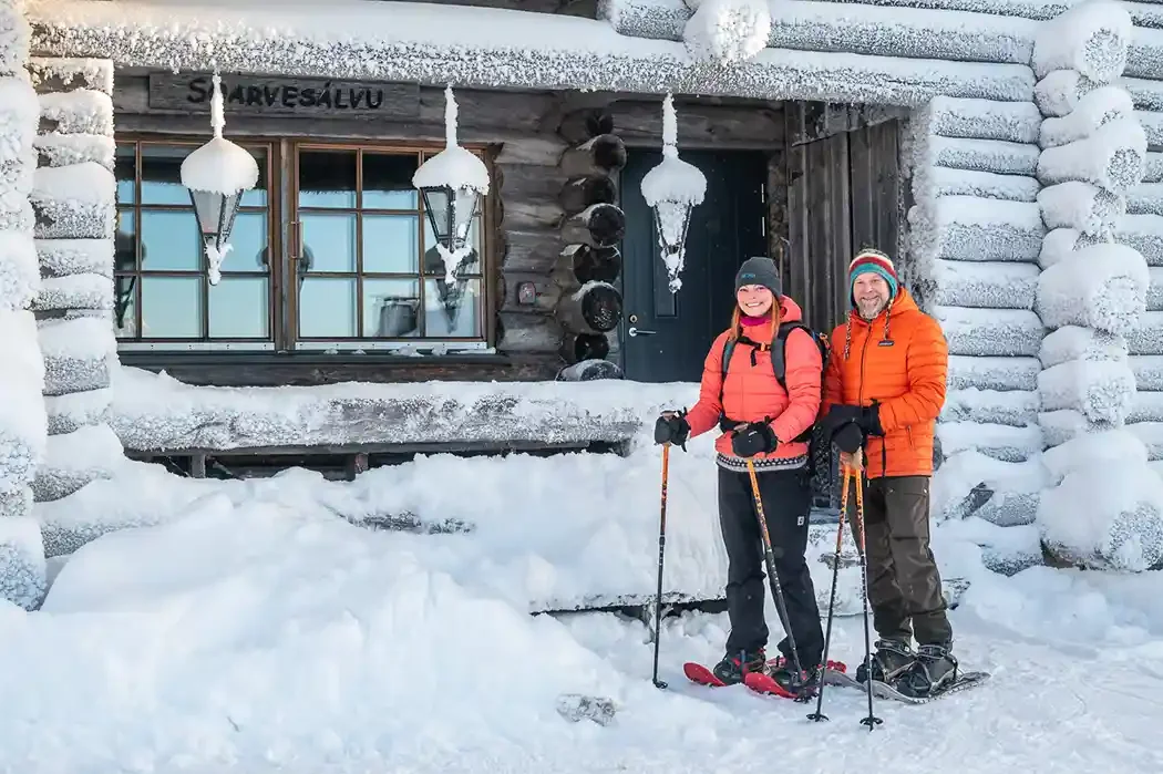 Two people in winter gear standing with ski poles in front of a snow-covered wooden cabin, smiling, with snow and ice on the cabin and windows.