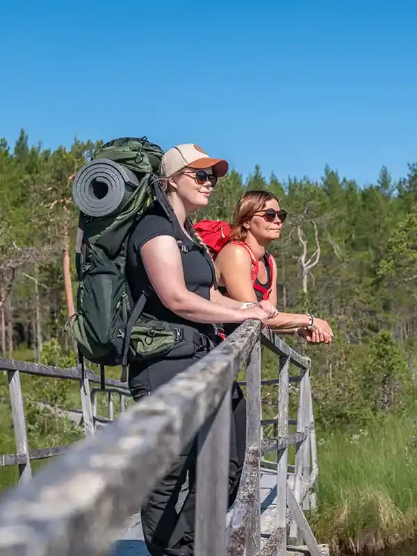 Two women with backpacks standing on a wooden bridge, overlooking a forested landscape under a clear blue sky.