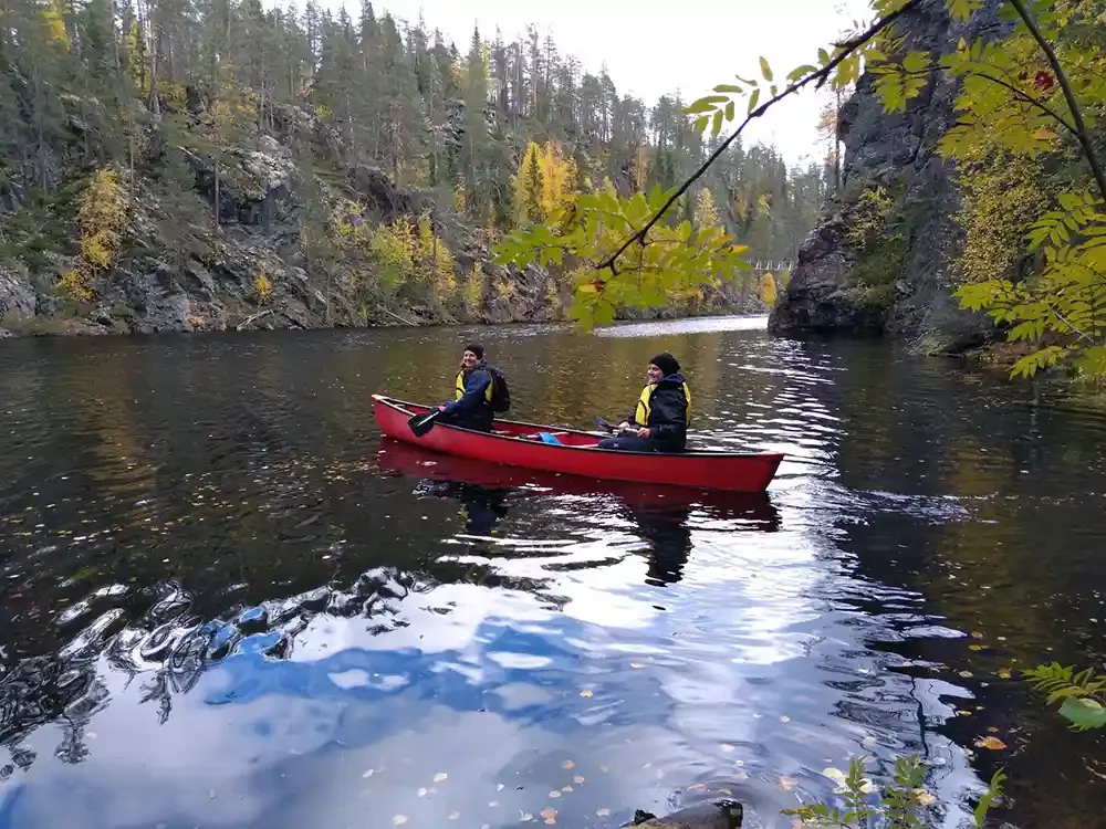 Two people kayaking on a calm river surrounded by rocky cliffs and trees with autumn foliage.