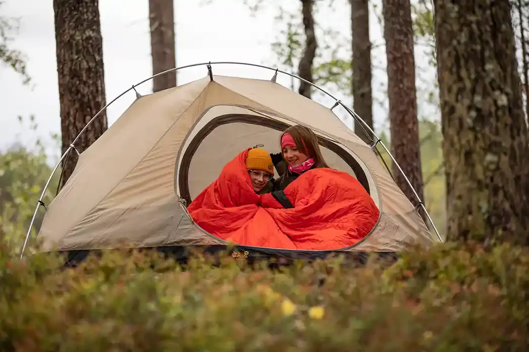 Two children inside a beige camping tent with orange sleeping bags in a forest with tall trees and green foliage.