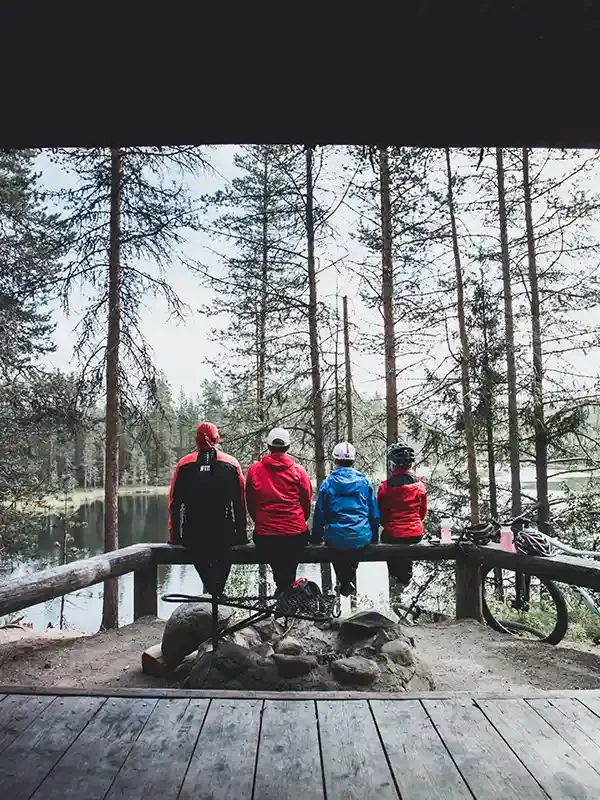 Four people sitting on a wooden railing overlooking a forested lake, with two bicycles nearby.