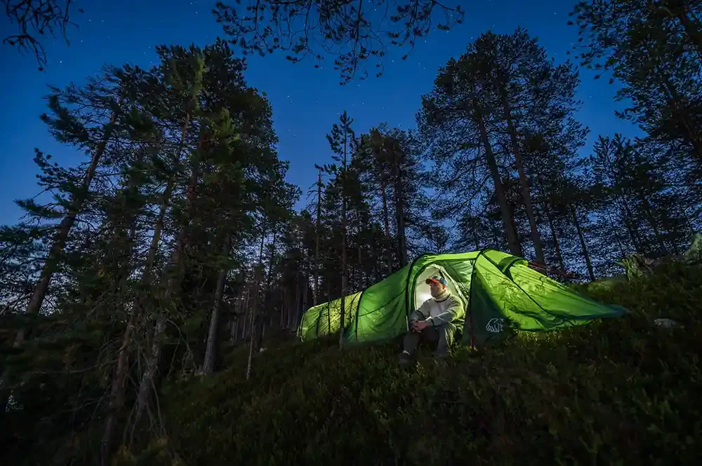 Person sitting outside a glowing green tent in a forest at dusk.
