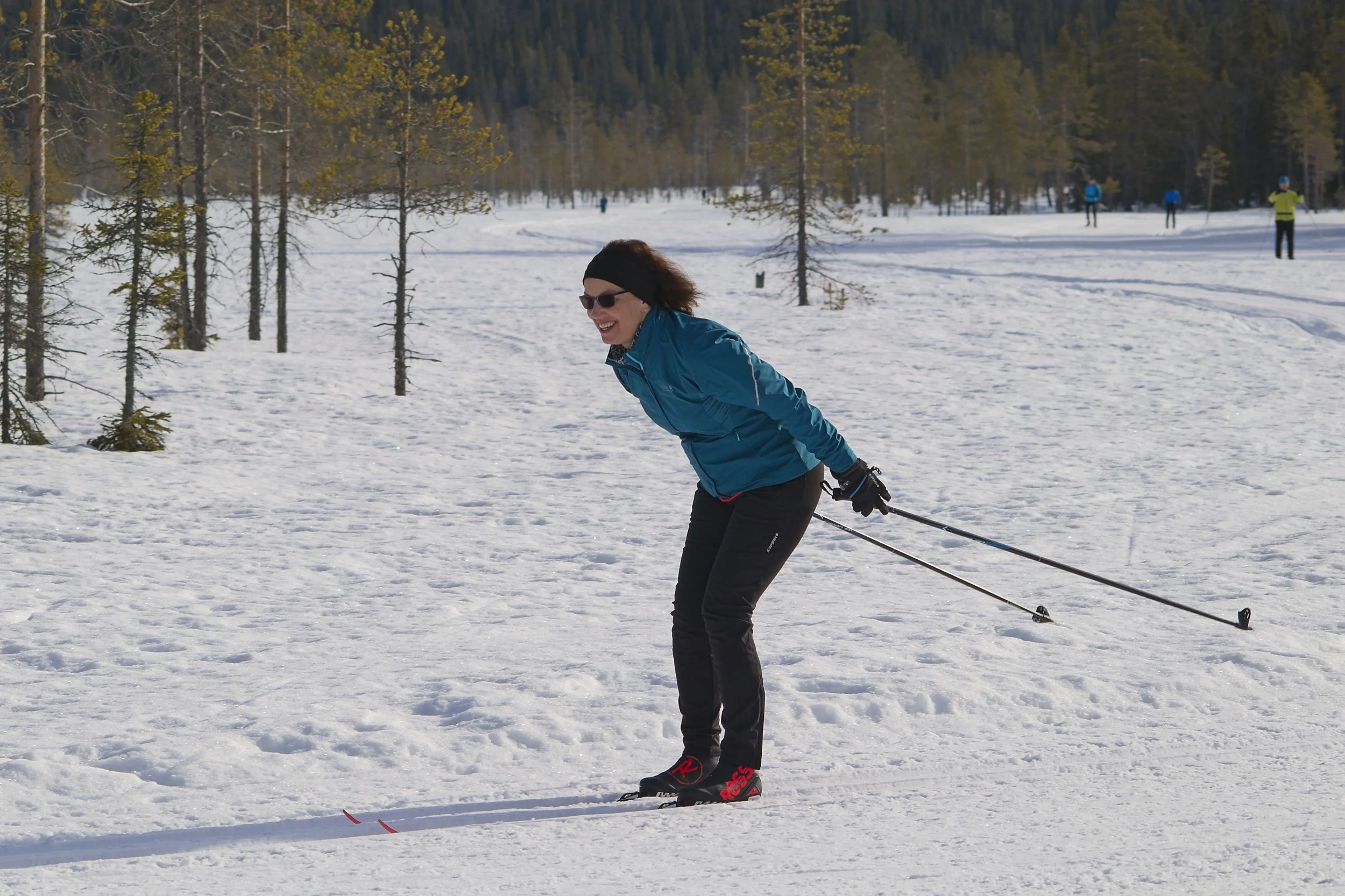 A woman in blue winter jacket, black headband, and sunglasses cross-country skiing in a snowy forest, with other skiers visible in the background.