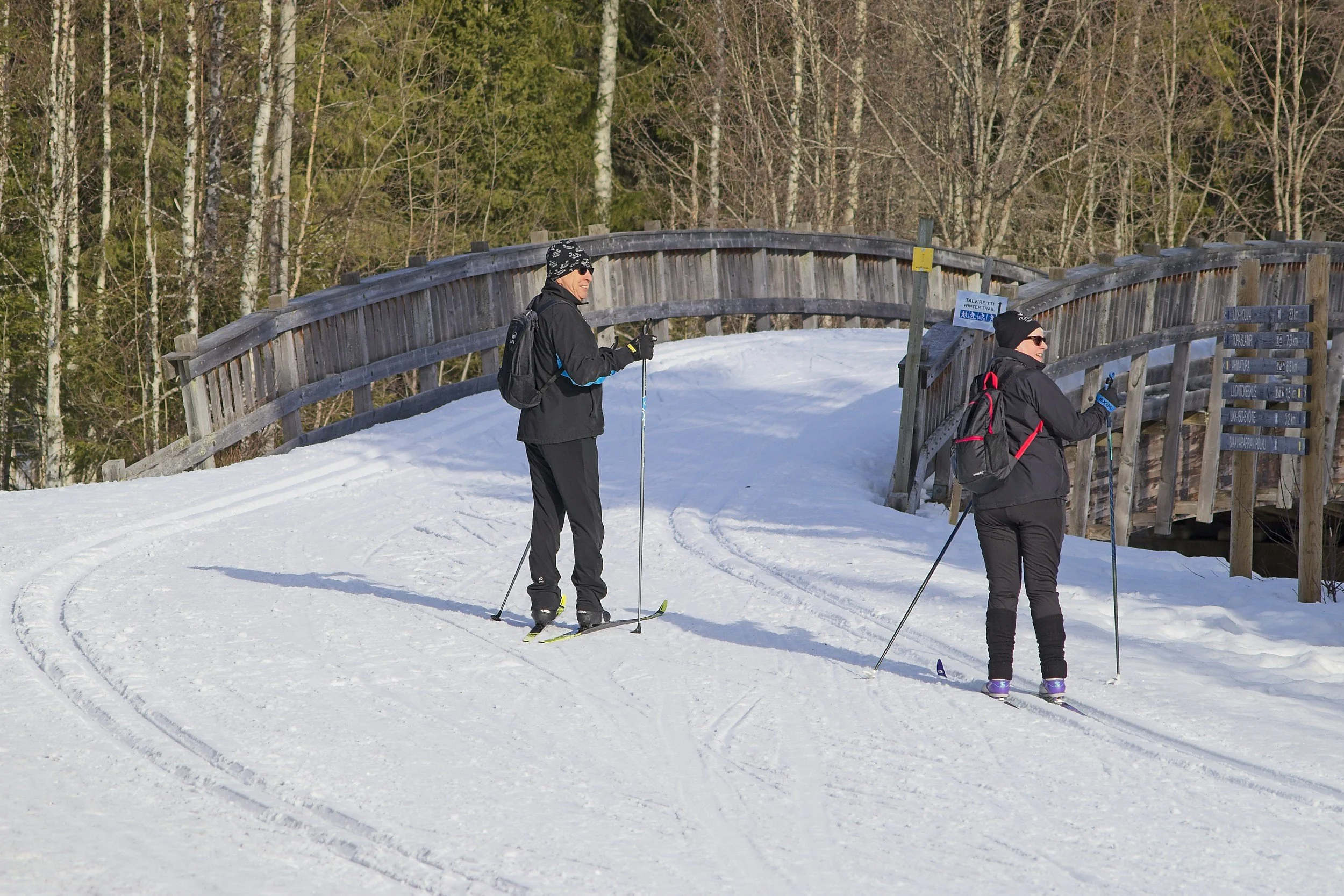Two people dressed in winter clothing, including hats and sunglasses, are cross-country skiing on a snow-covered trail in a forested area during daytime. They are standing near a wooden trail sign and a bridge.