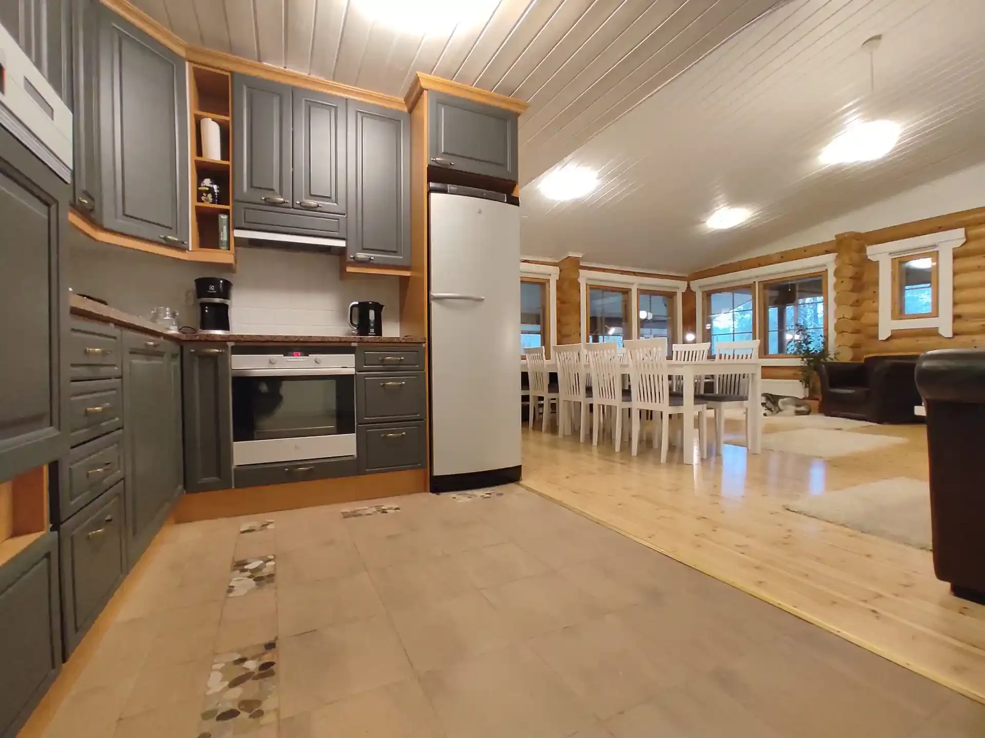 Interior of a log cabin with a view of the kitchen and dining area, featuring gray cabinets, a white refrigerator, a dining table with white chairs, and a cozy living space with black couches and large windows.