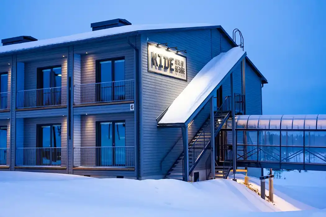 A modern two-story building with a sign that reads 'KIDS' and 'Outdoor & Indoor Play Space', surrounded by snow and featuring an external staircase and a glass-enclosed walkway connecting to another structure.