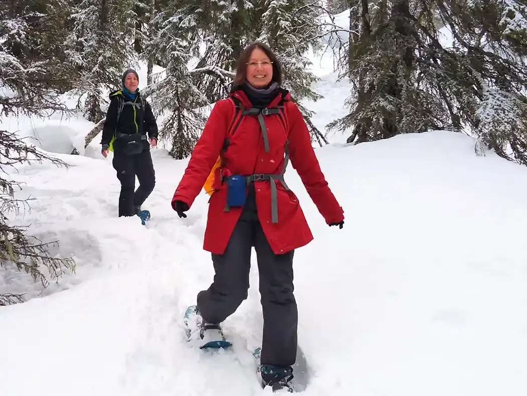 Two women hiking in snowy forest with snowshoes, wearing winter gear and backpacks.