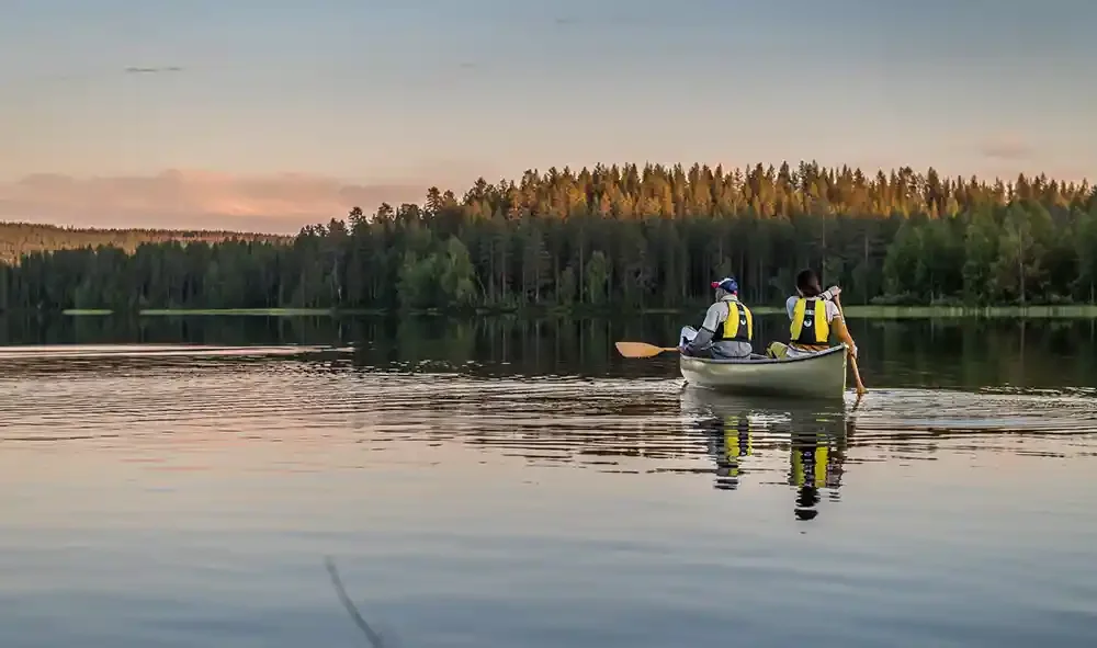 Two people in a canoe on a calm lake during sunset, surrounded by a forested landscape.