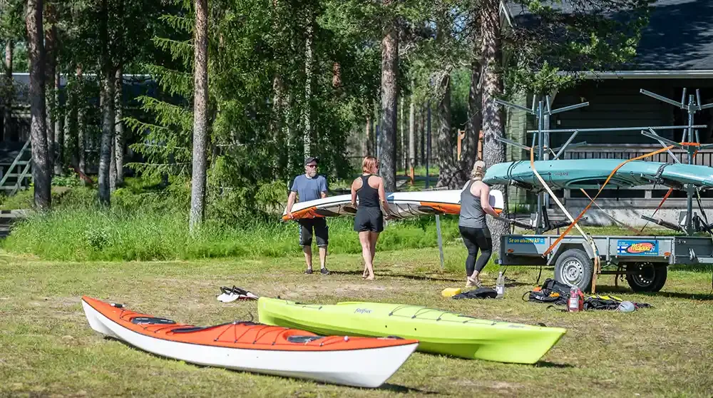 People preparing kayaks and canoes at a lakeside or riverside area surrounded by trees, with equipment on a trailer.