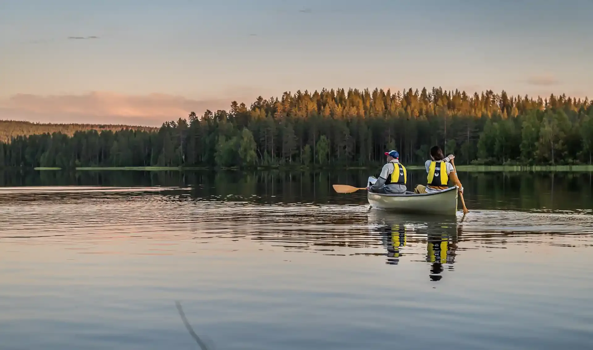 Two people in a canoe paddling on a calm lake during sunset, with a dense forest on the shoreline in the background.