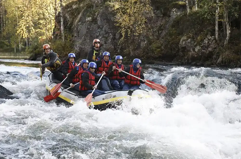 Group of people white water rafting on a river with rocky shoreline and trees in the background.