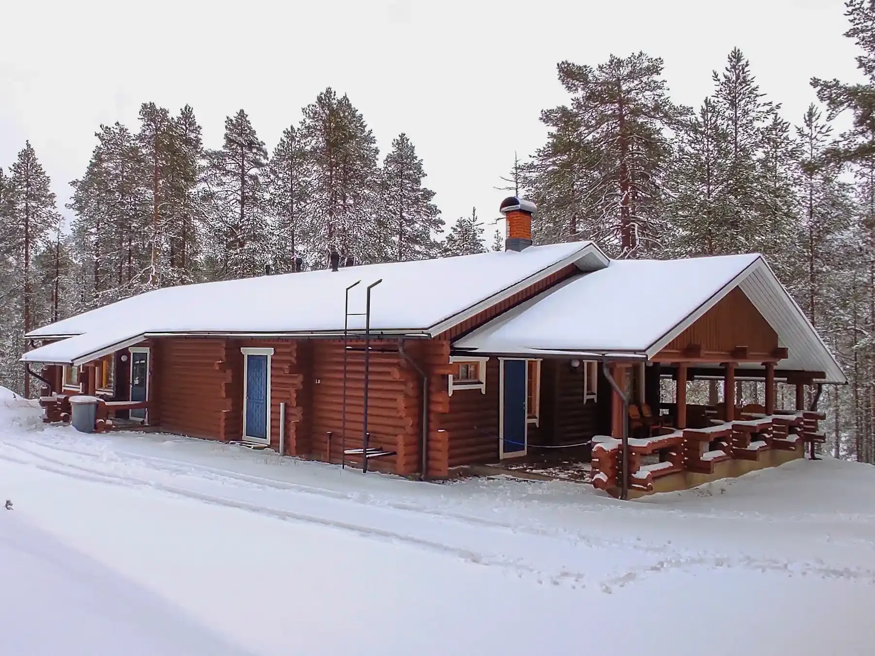 A wooden house with a snow-covered sloped roof in a forested area during winter, surrounded by tall pine trees.