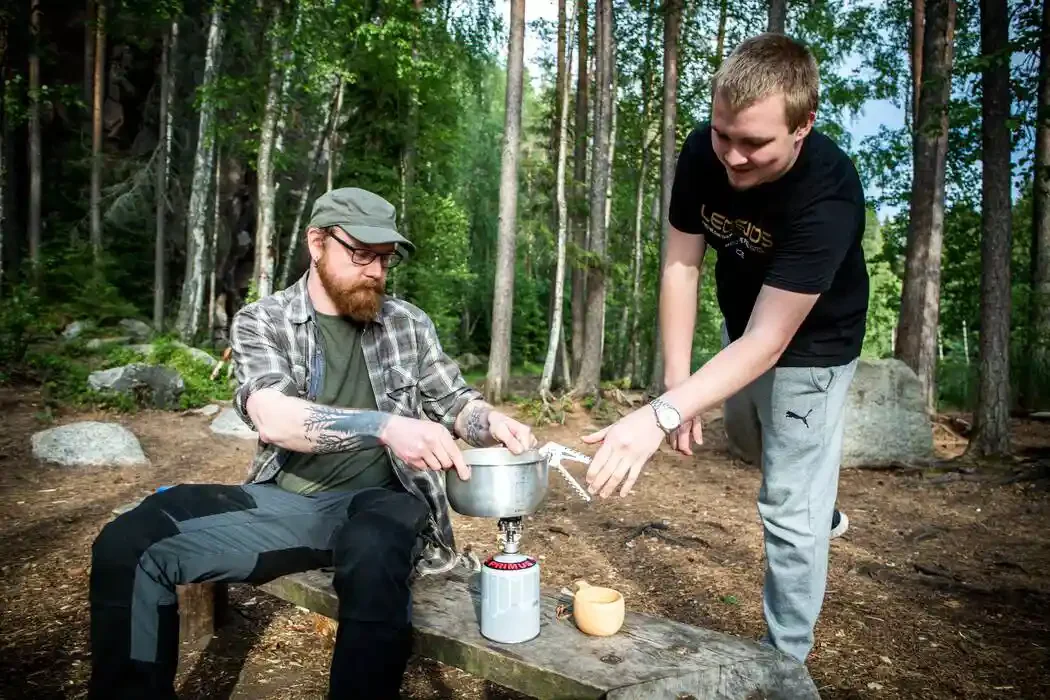Two men cooking outdoors in a forest, one sitting on a log holding a pot over a portable stove, the other leaning over, assisting with the pot.