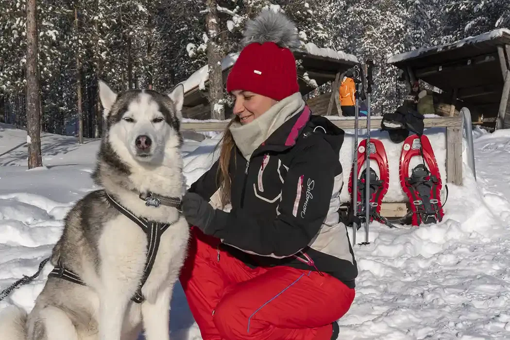 A woman in winter clothing sitting next to a Siberian Husky dog in a snowy outdoor setting, with snowshoes and a wooden shelter in the background.