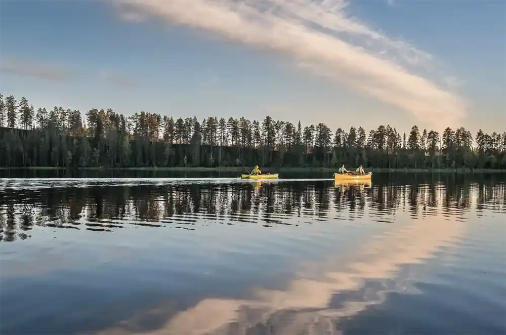 Two people in yellow canoes on a calm lake surrounded by trees, with a partly cloudy sky reflecting on the water.