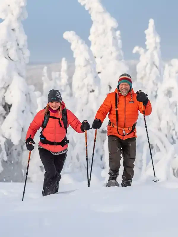 A smiling man and woman hiking through snowy landscape with snow-covered trees in the background, dressed in orange jackets, black gloves, hats, and using hiking poles.