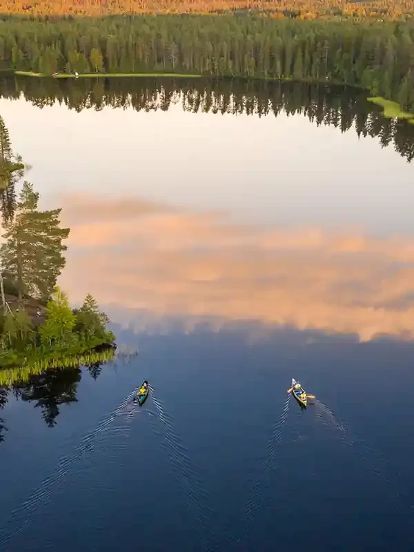 Two kayakers paddle on a calm lake surrounded by trees, with a reflection of the sky and clouds on the water.