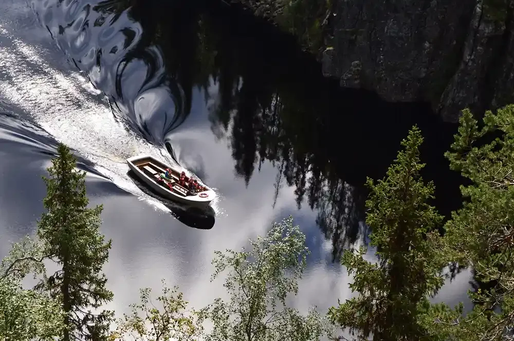 A small boat with people on it sailing through a dark, calm river surrounded by trees and rocky cliffs.