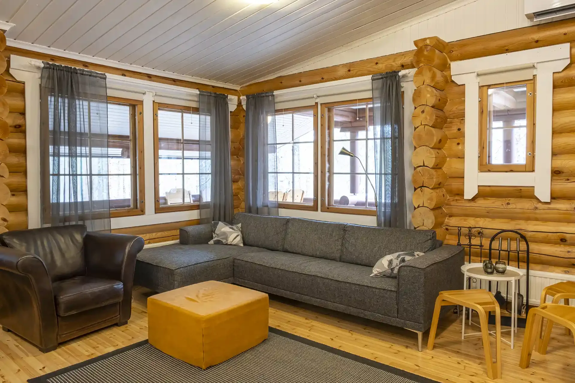 Living room with log cabin interior, large gray sectional sofa, black leather armchair, wooden coffee table, windows with sheer curtains, and a small side table with decorative items.