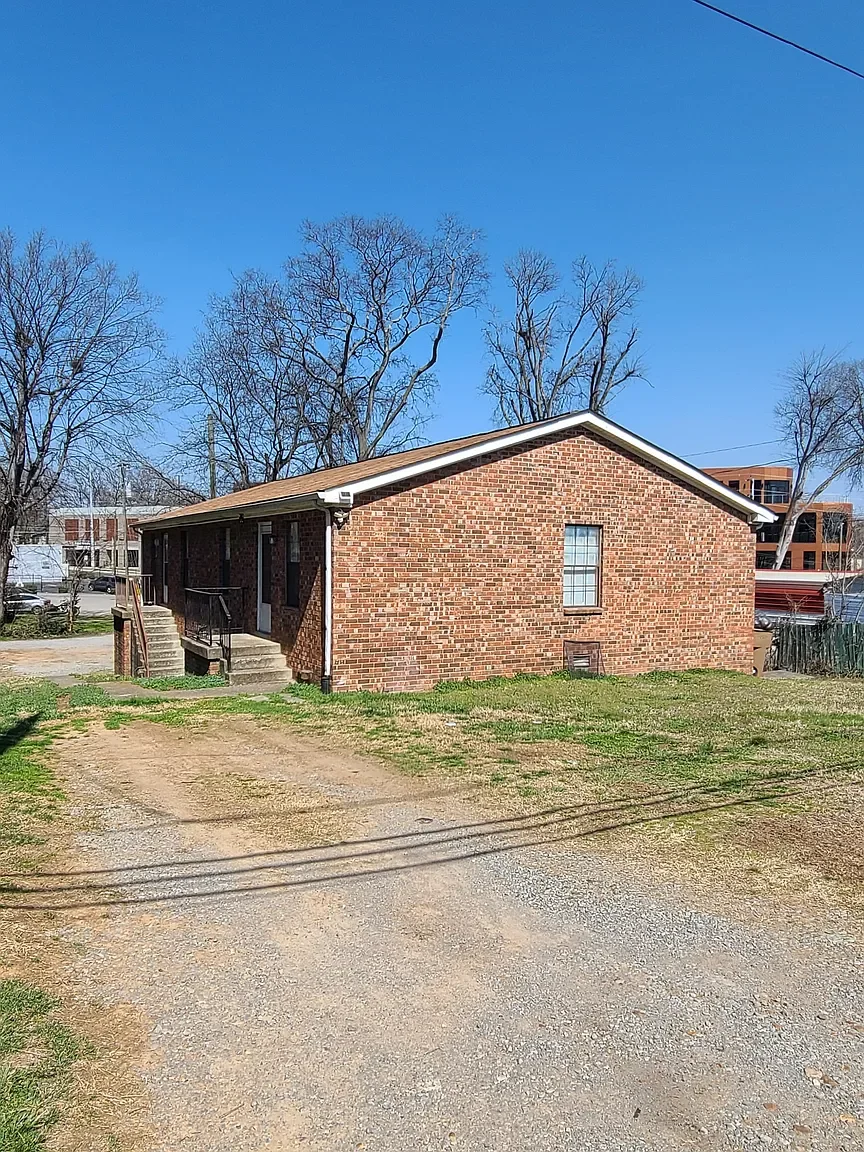 A single-story brick house with a sloped roof, steps leading to a side entrance, and a grassy yard with a dirt driveway. Tall trees with bare branches are in the background under a clear blue sky.